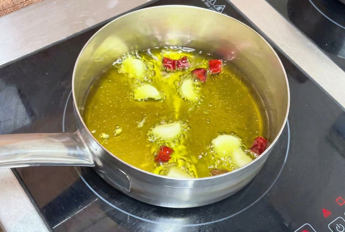 Ząbki czosnku i chili smażące się w bulgoczącej oliwie w małym metalowym garnku na kuchence.