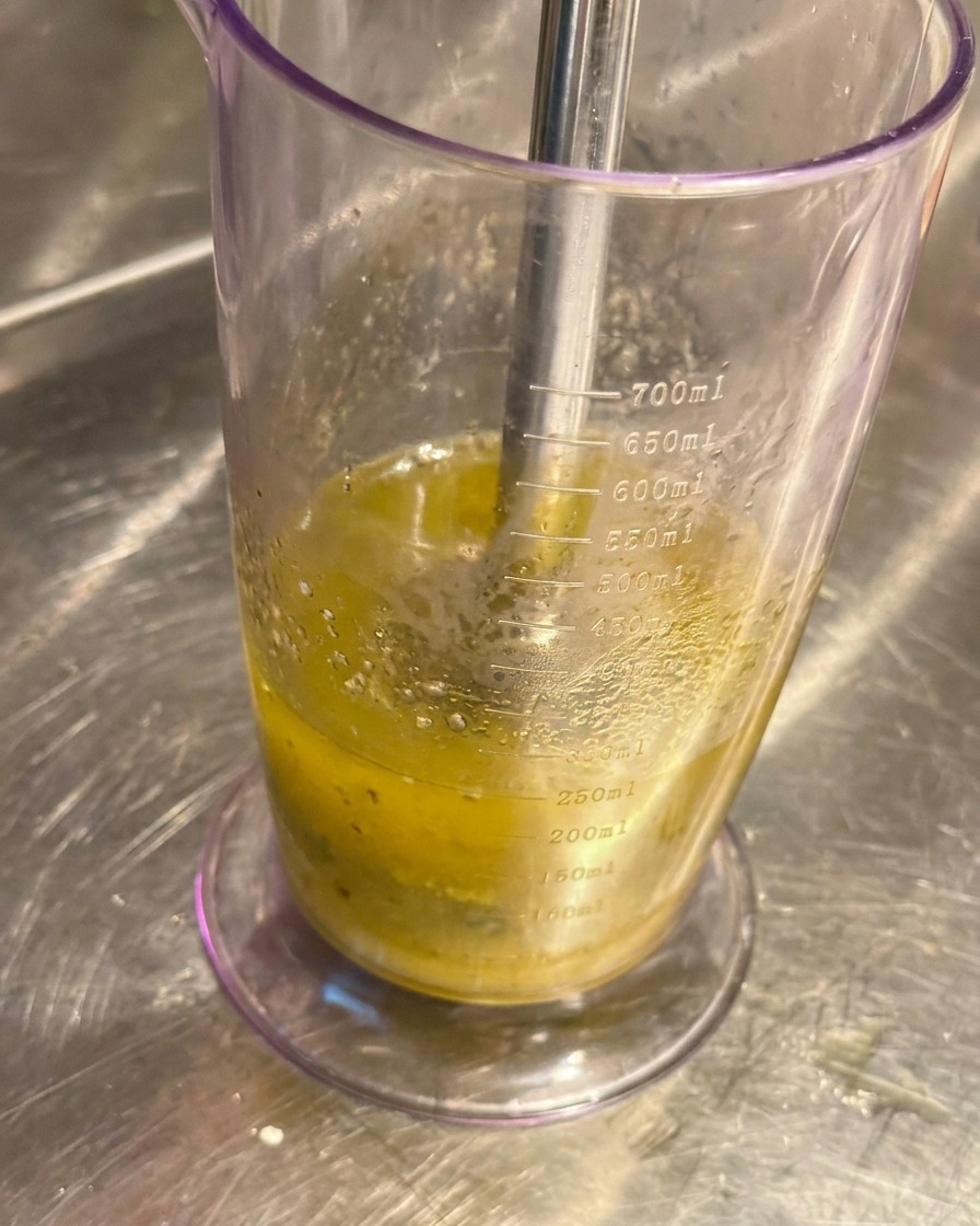 Blender ręczny zanurzony w wysokim naczyniu, miksujący czosnek z oliwą na jasną pastę.