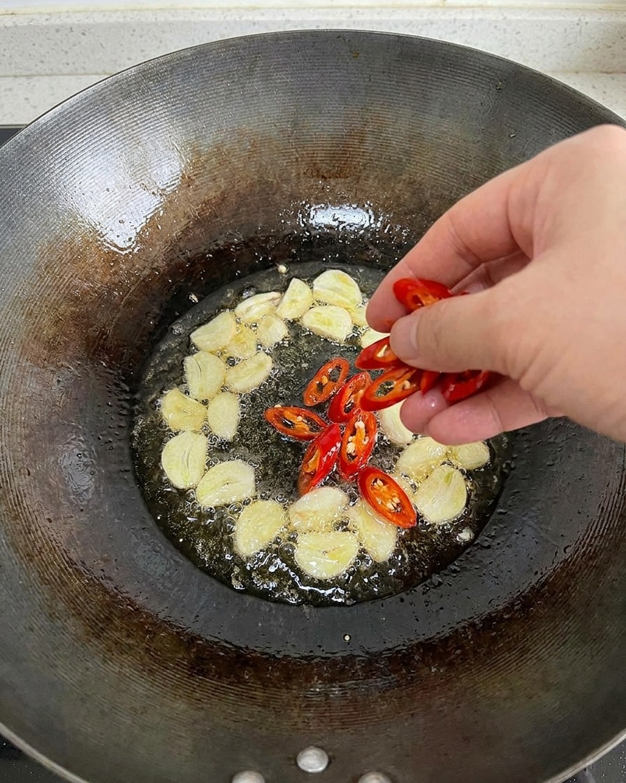 Ręka wrzucająca świeżo posiekane papryczki chili do woka z gorącą oliwą, w której już smażą się plastry czosnku.