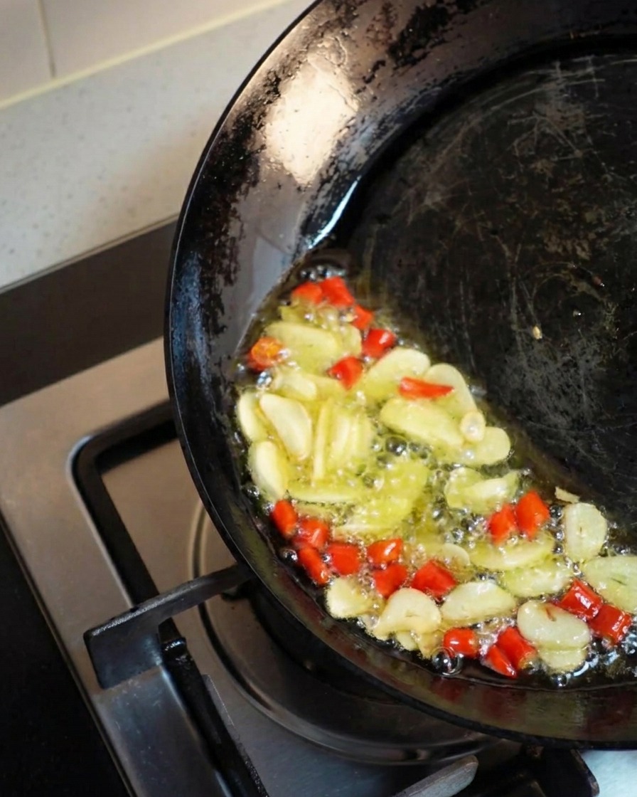 Ciemny wok przechylony na bok, w którym złocista oliwa z czosnkiem i chili intensywnie skwierczy i smaży się razem.