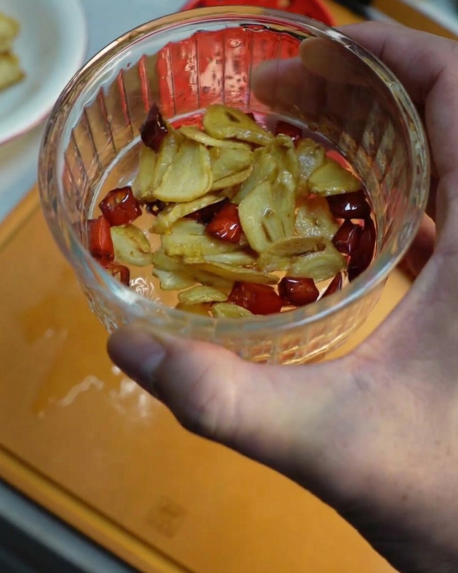 Ręka trzymająca małą szklaną miseczkę wypełnioną chrupiącymi plastrami smażonego czosnku i kawałkami czerwonego chili.