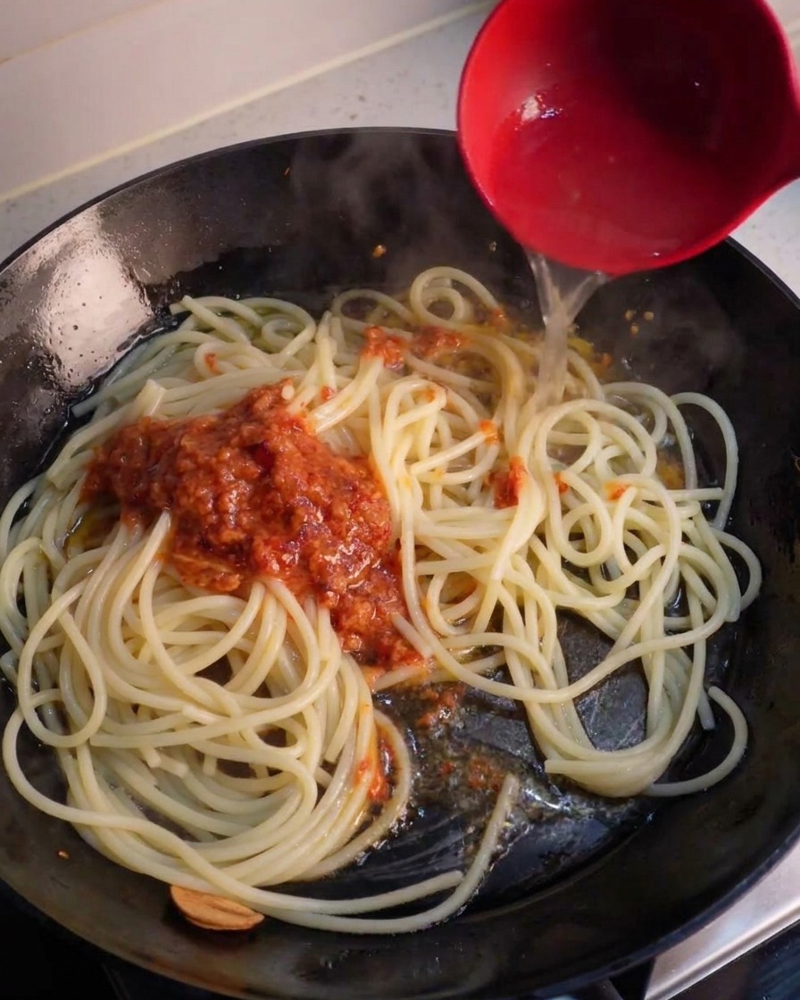 Czerwona chochla wlewająca wodę z makaronu na patelnię ze spaghetti i porcją zmiksowanego sosu chili.