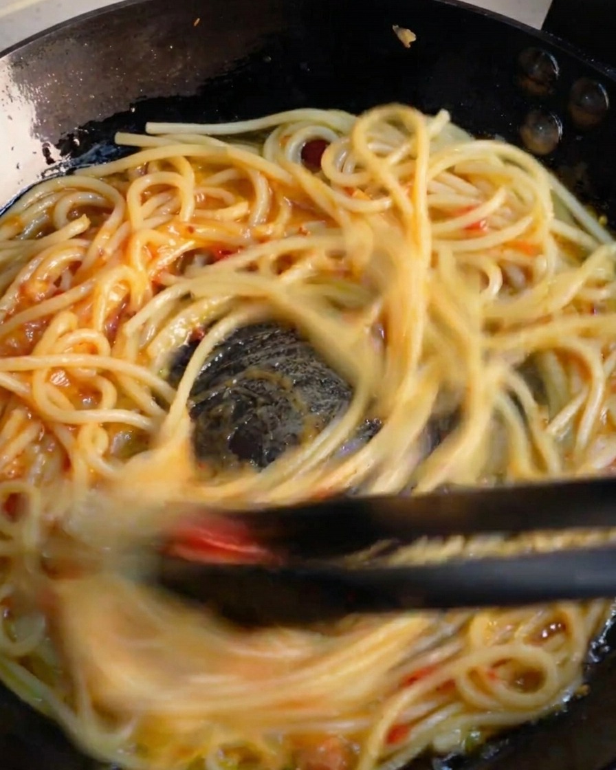 Czarne szczypce kuchenne szybko mieszające spaghetti na patelni, aby połączyć składniki w intensywny sos.
