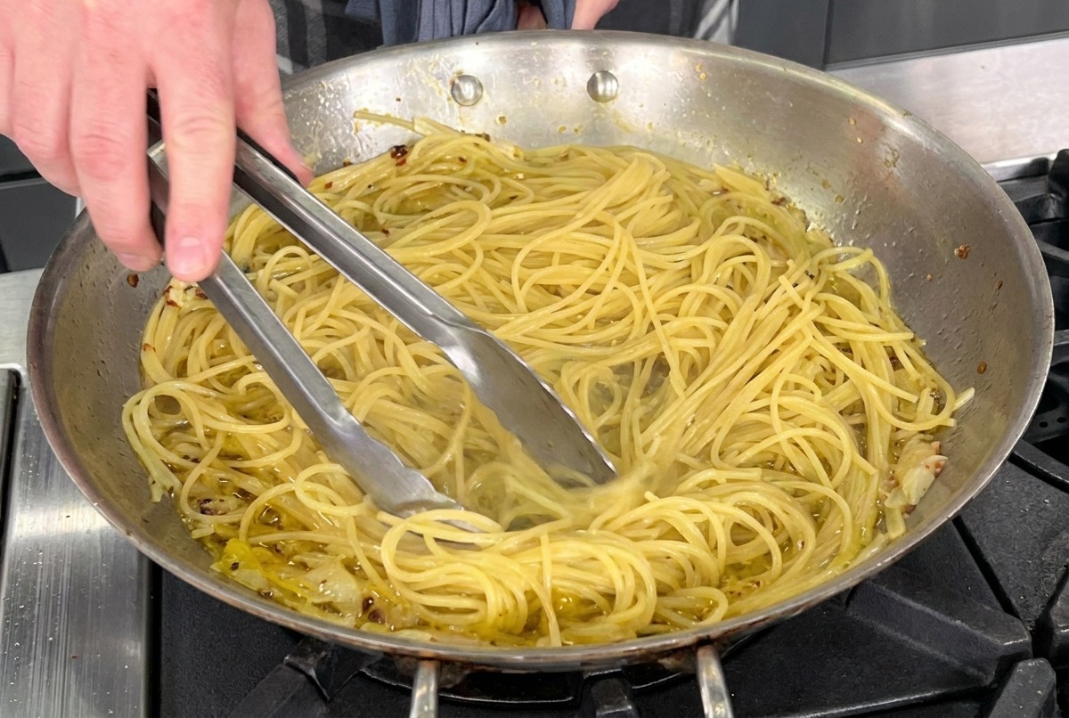 Szczypce mieszające makaron z oliwą, czosnkiem i chili na stalowej patelni.
