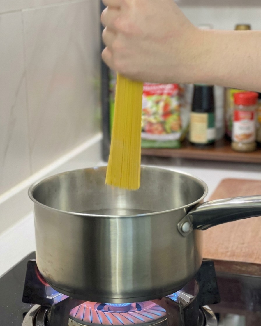 Dłoń wrzucająca pionowo garść suchego spaghetti do garnka ze stali nierdzewnej z wrzącą wodą.