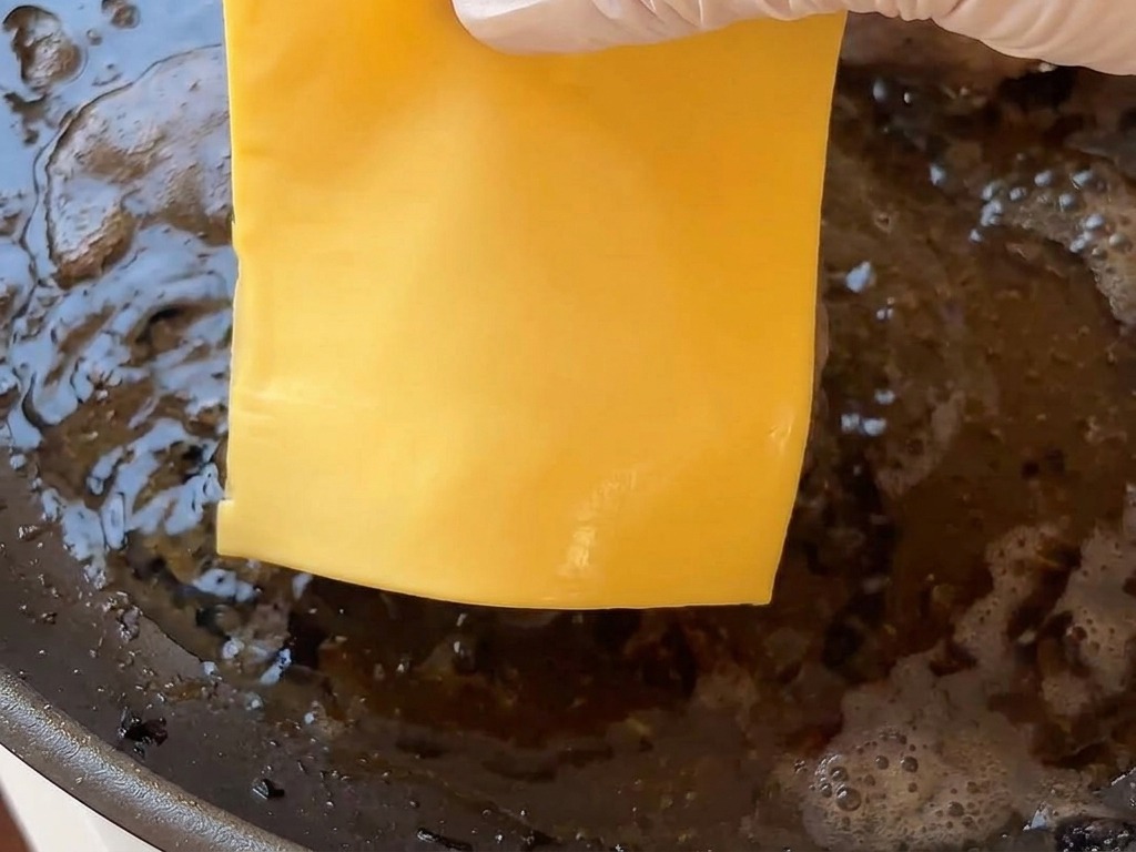 Dłoń nakładająca plaster sera na gorący kotlet wołowy smażący się na patelni.
