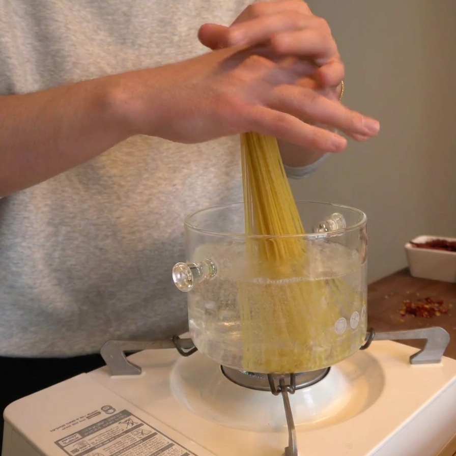 Dłonie wrzucające garść suchego spaghetti pionowo do szklanego garnka z wrzącą wodą.