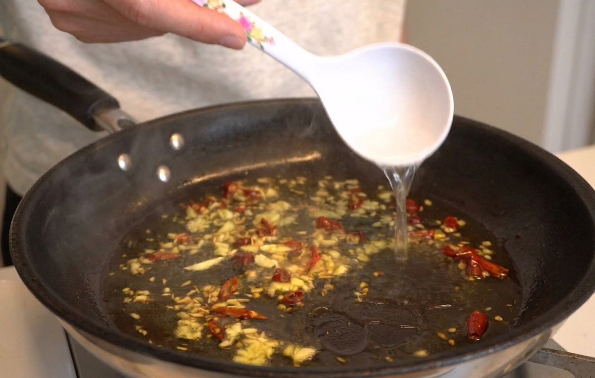 Biała chochla wlewająca gorącą, skrobiową wodę na patelnię ze skwierczącym czosnkiem, oliwą i chili.