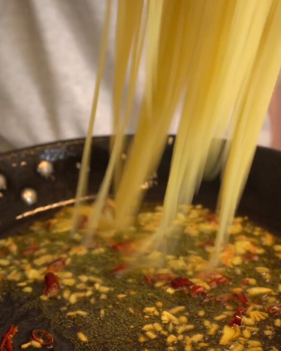 Długie nitki spaghetti przenoszone na patelnię z powoli gotującym się sosem czosnkowo-paprykowym.