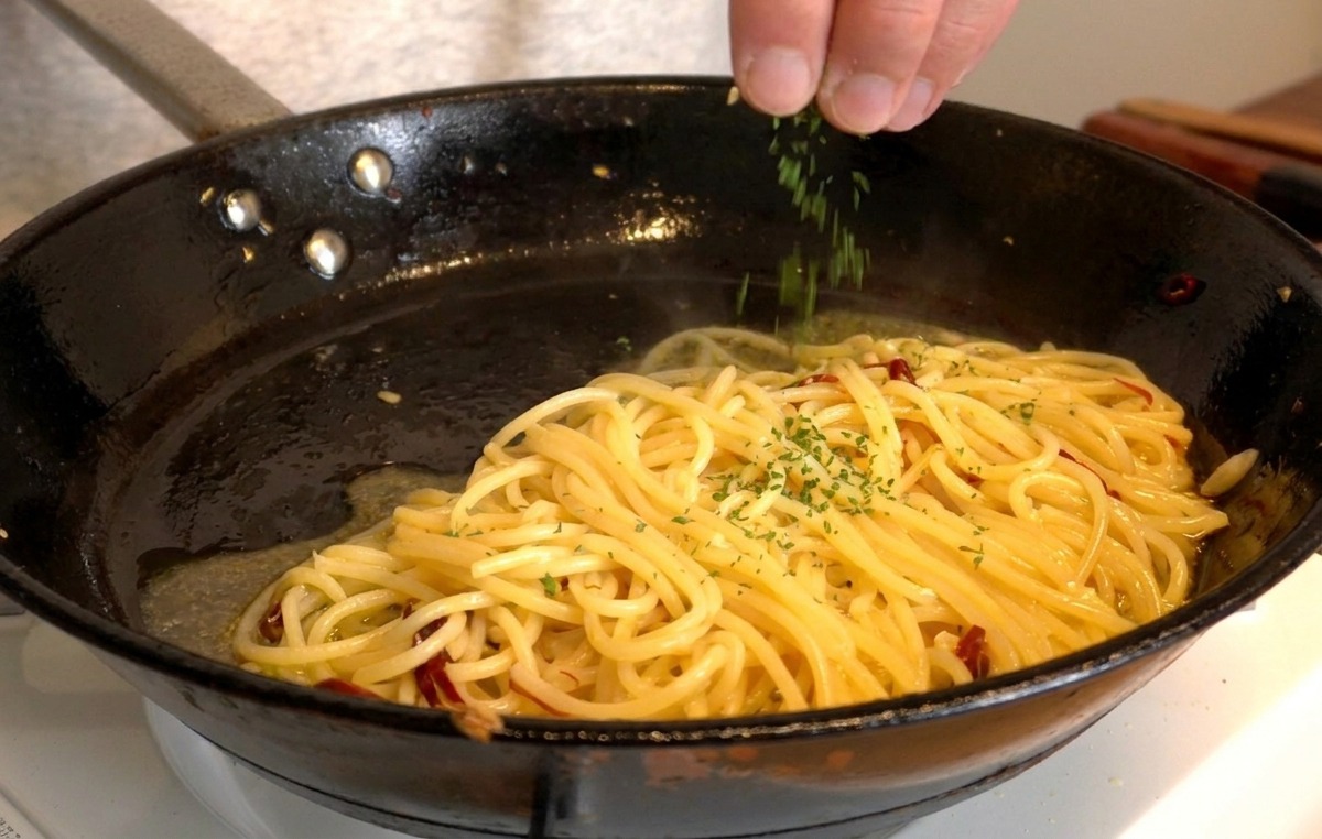 Dłoń posypująca posiekaną, świeżą natką pietruszki lśniące od oliwy spaghetti na patelni.