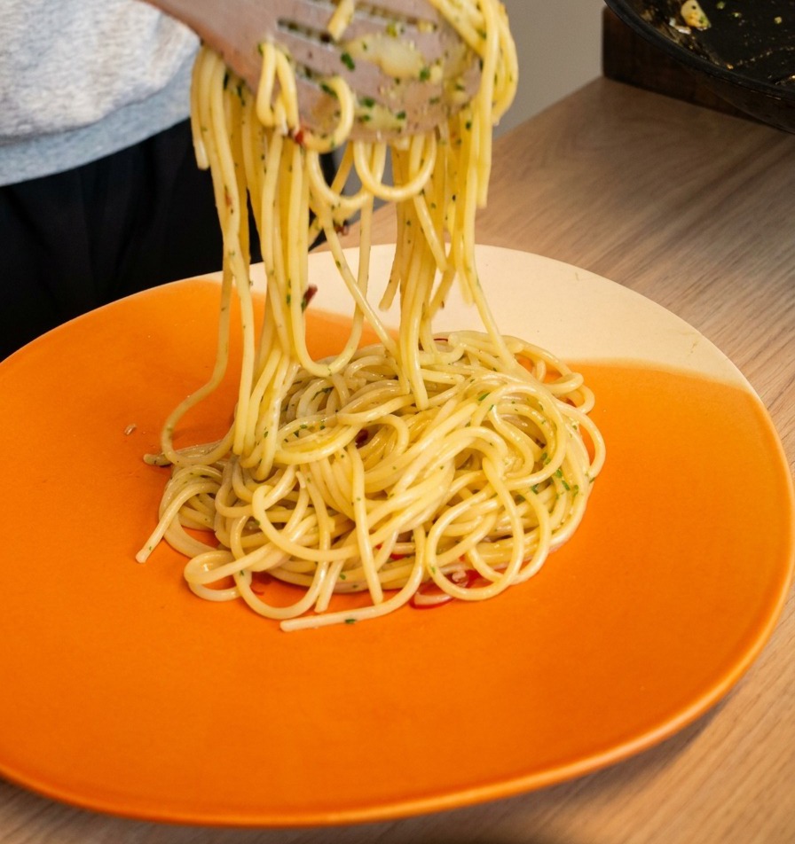 Szczypce unoszące i owijające porcję spaghetti aglio e olio na jasnopomarańczowym talerzu.