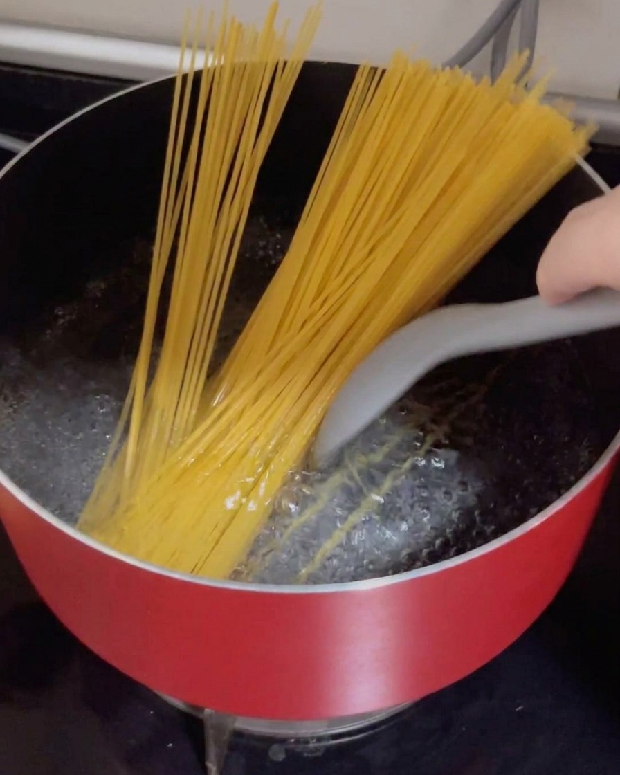 Suche nitki spaghetti wkładane do czerwonego garnka z mocno wrzącą wodą.