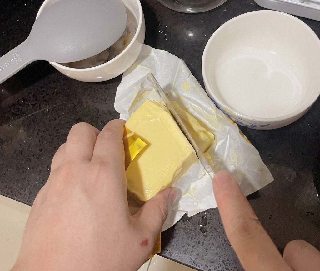 Osoba odkrawająca gruby plaster masła z kostki za pomocą dużego noża kuchennego.