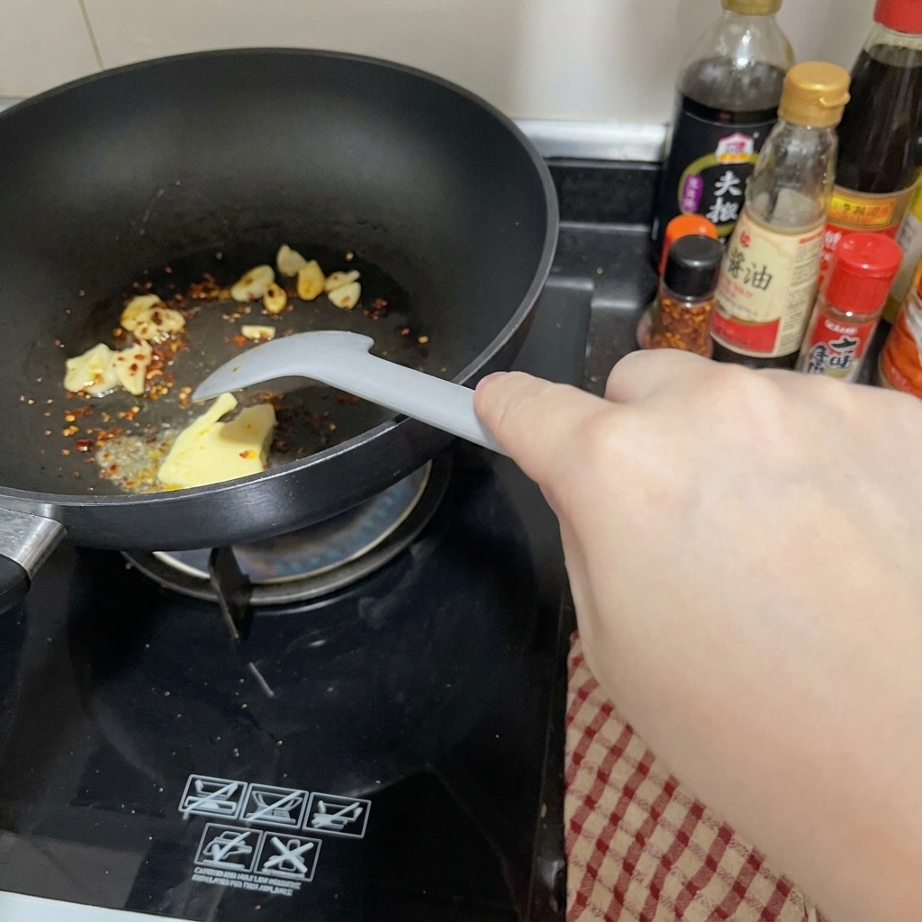 Kawałek masła roztapiający się na czarnej patelni obok czosnku i chili, mieszany szarą łopatką.