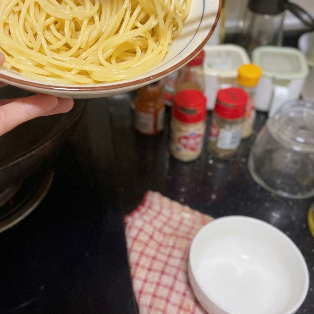 Dłoń trzymająca miskę z ugotowanym makaronem nad patelnią, przygotowując się do jego dodania.