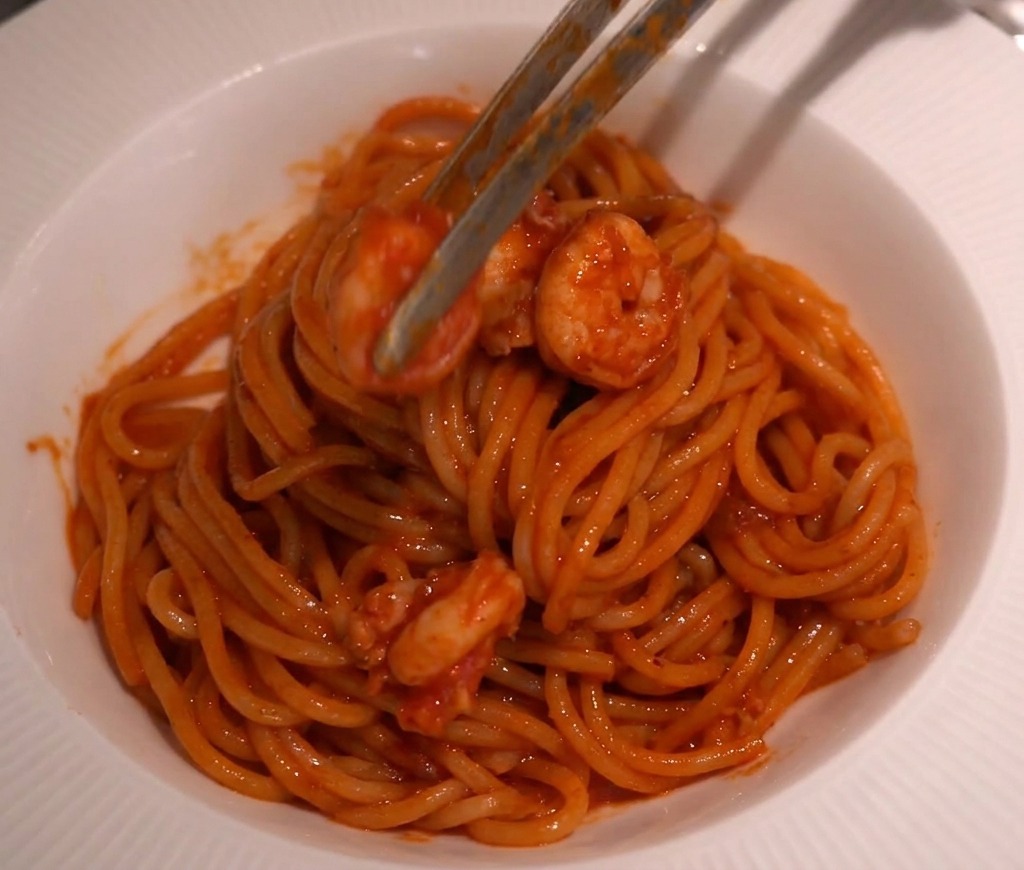 Szczypce kuchenne ostrożnie układające krewetki w sosie na szczycie zawiniętego gniazdka ze spaghetti w białej misce.
