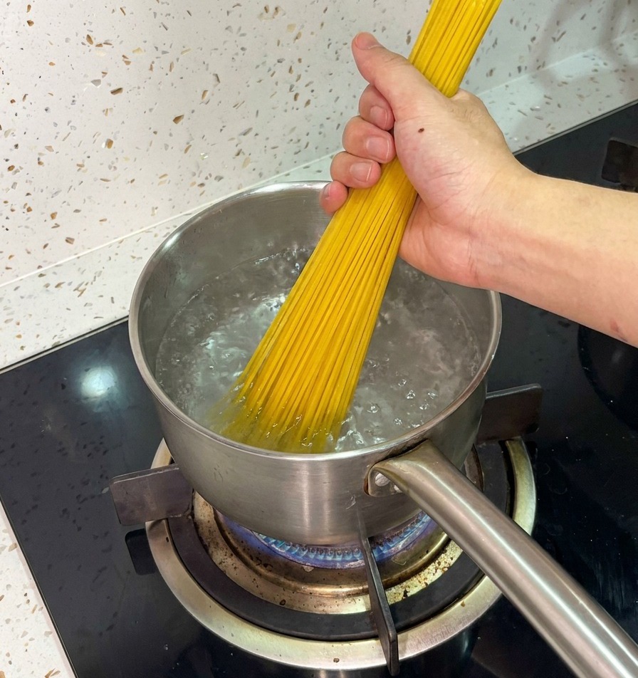 Dłoń trzymająca pęczek suchego spaghetti, wkładająca go do garnka z wrzącą wodą.