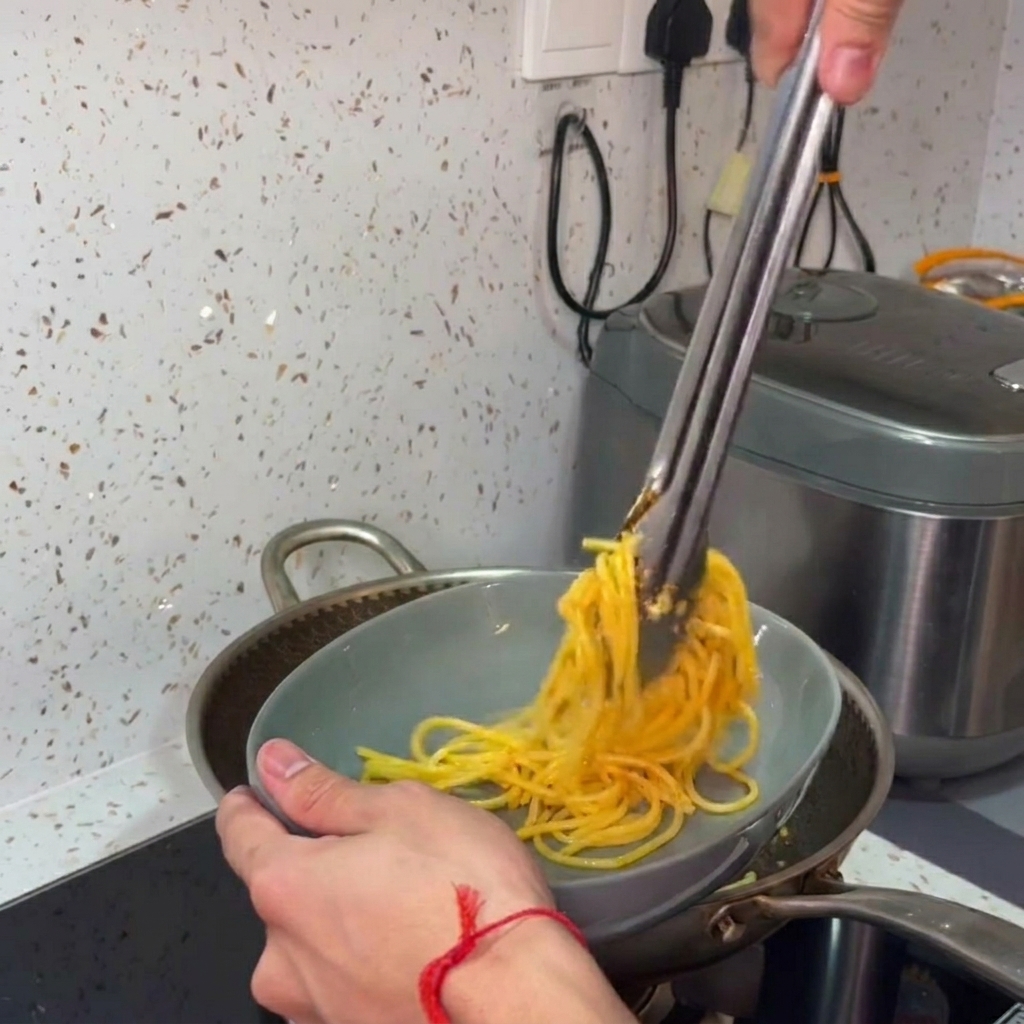 Dłonie nakładające i zwijające gorące spaghetti szczypcami z patelni do szarej miski.