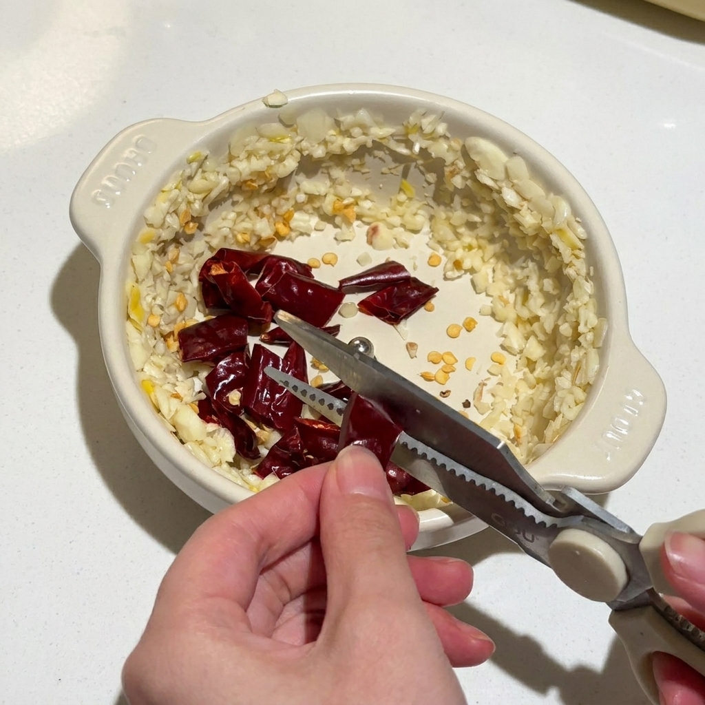 Ręce używające nożyczek kuchennych do cięcia suszonych papryczek chili do miski z posiekanym czosnkiem.