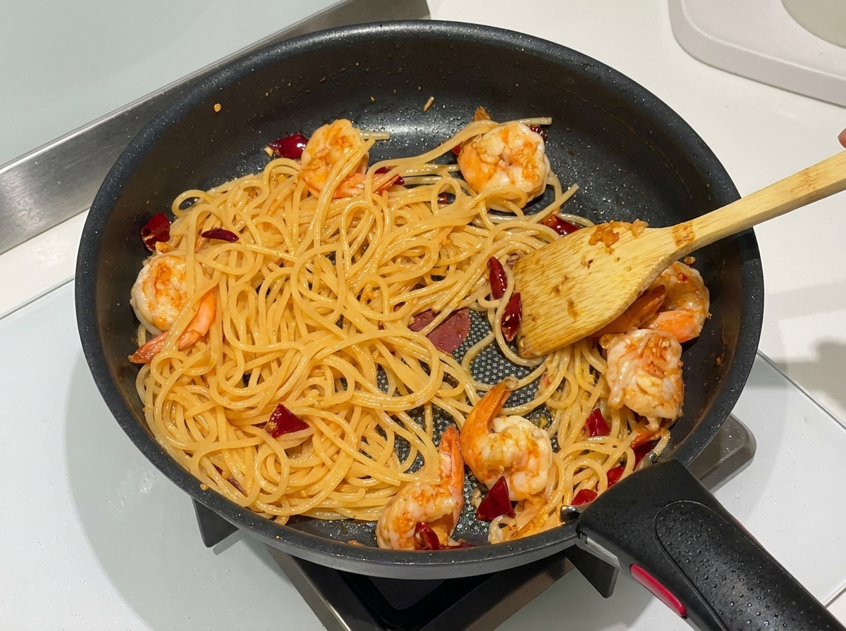 Ugotowane spaghetti, różowe krewetki i suszone chili mieszane razem na czarnej patelni drewnianą łopatką.