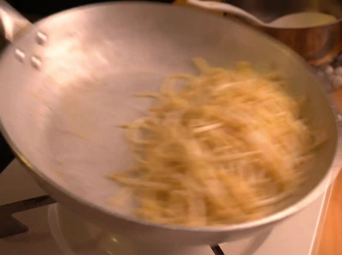 Energiczne mieszanie spaghetti na srebrnej patelni, widoczny efekt ruchu i emulgowania sosu.