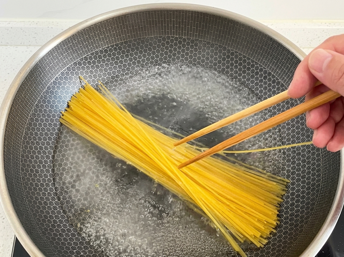 Dłoń używająca drewnianych pałeczek do zanurzania makaronu spaghetti w woku z gotującą się wodą.