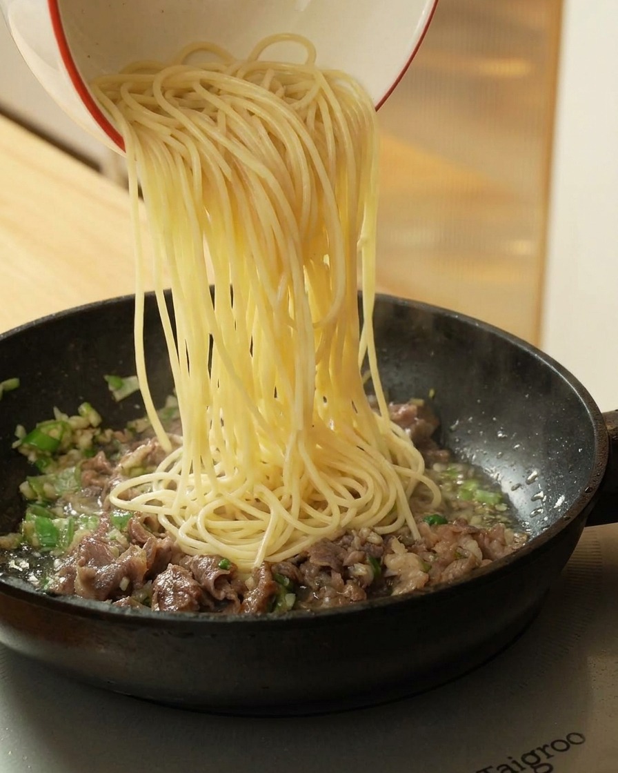 Przekładanie ugotowanego spaghetti z miski na patelnię z usmażoną wołowiną i aromatami.