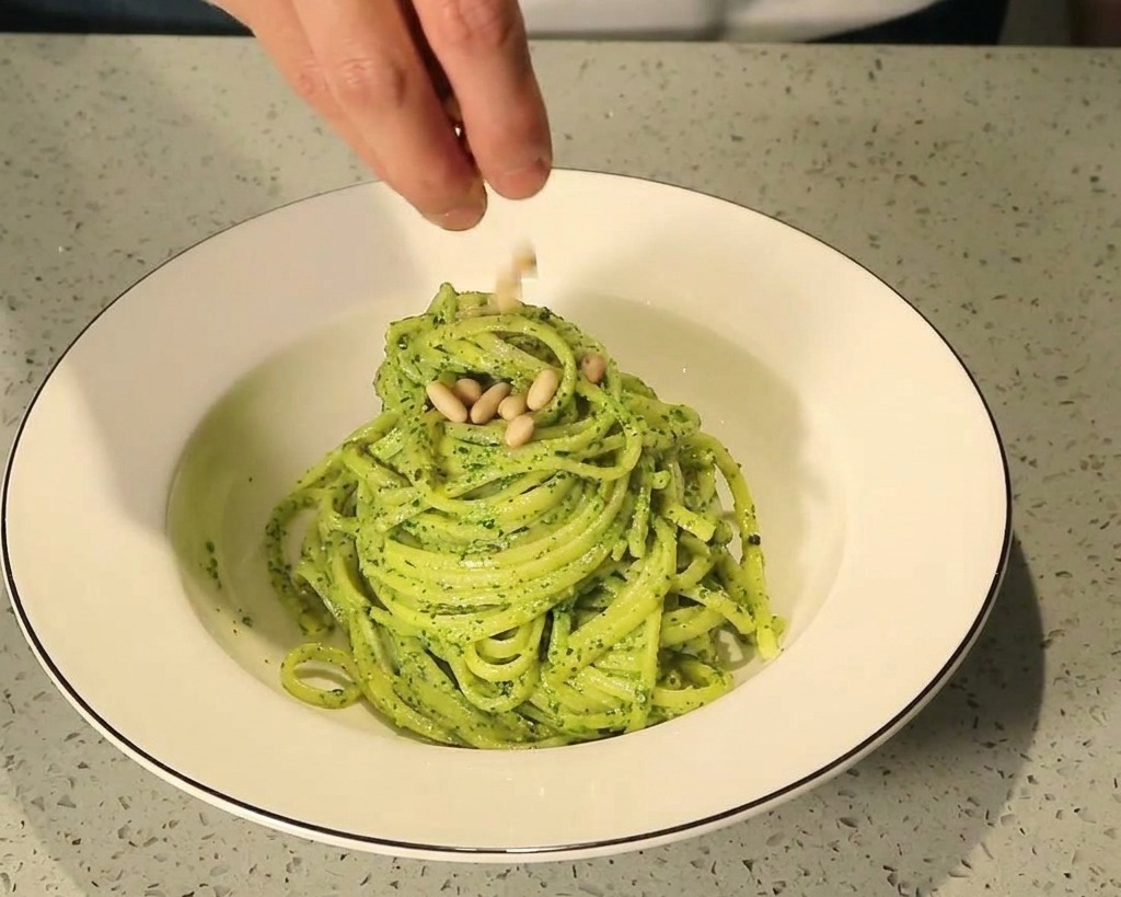 Palce posypujące prażonymi orzeszkami piniowymi kopczyk makaronu pesto w białej misce.