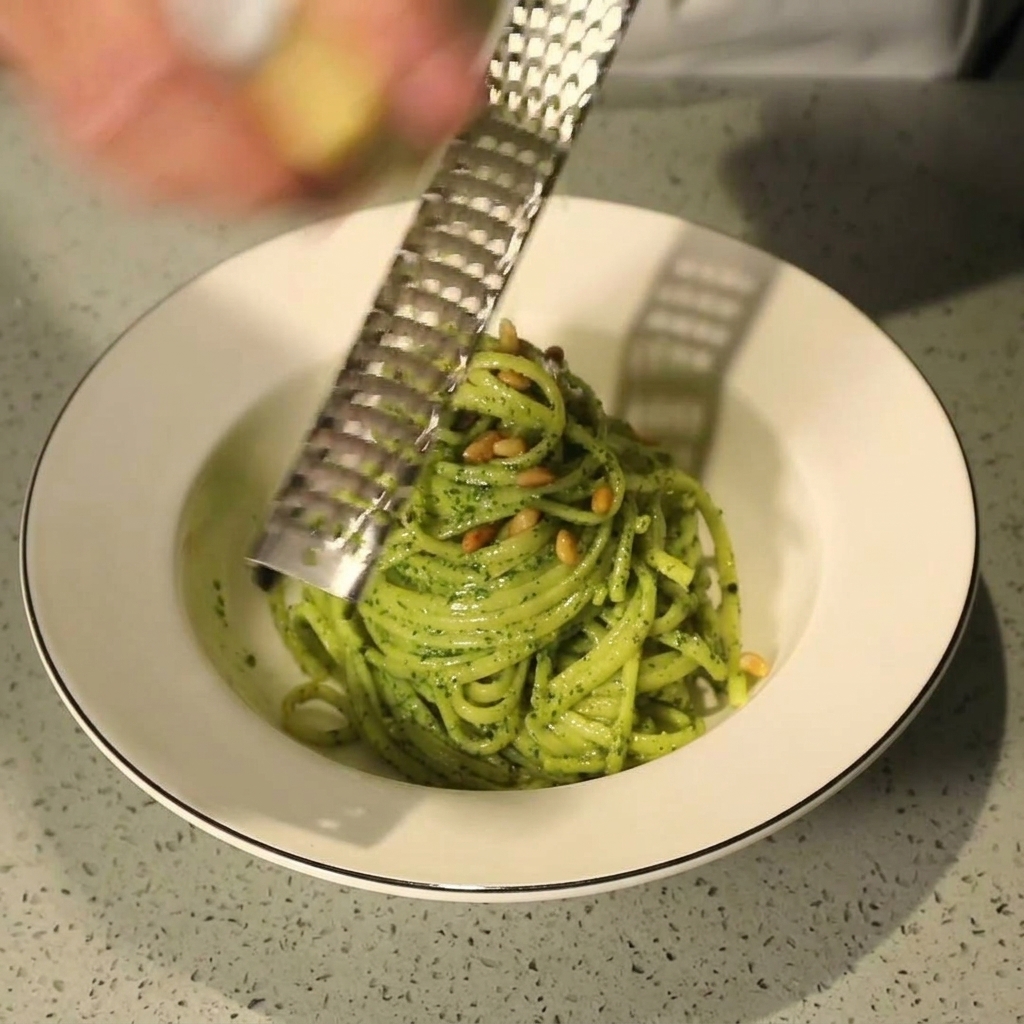 Ręka używająca metalowej tarki do ścierania świeżego parmezanu nad miską makaronu pesto.