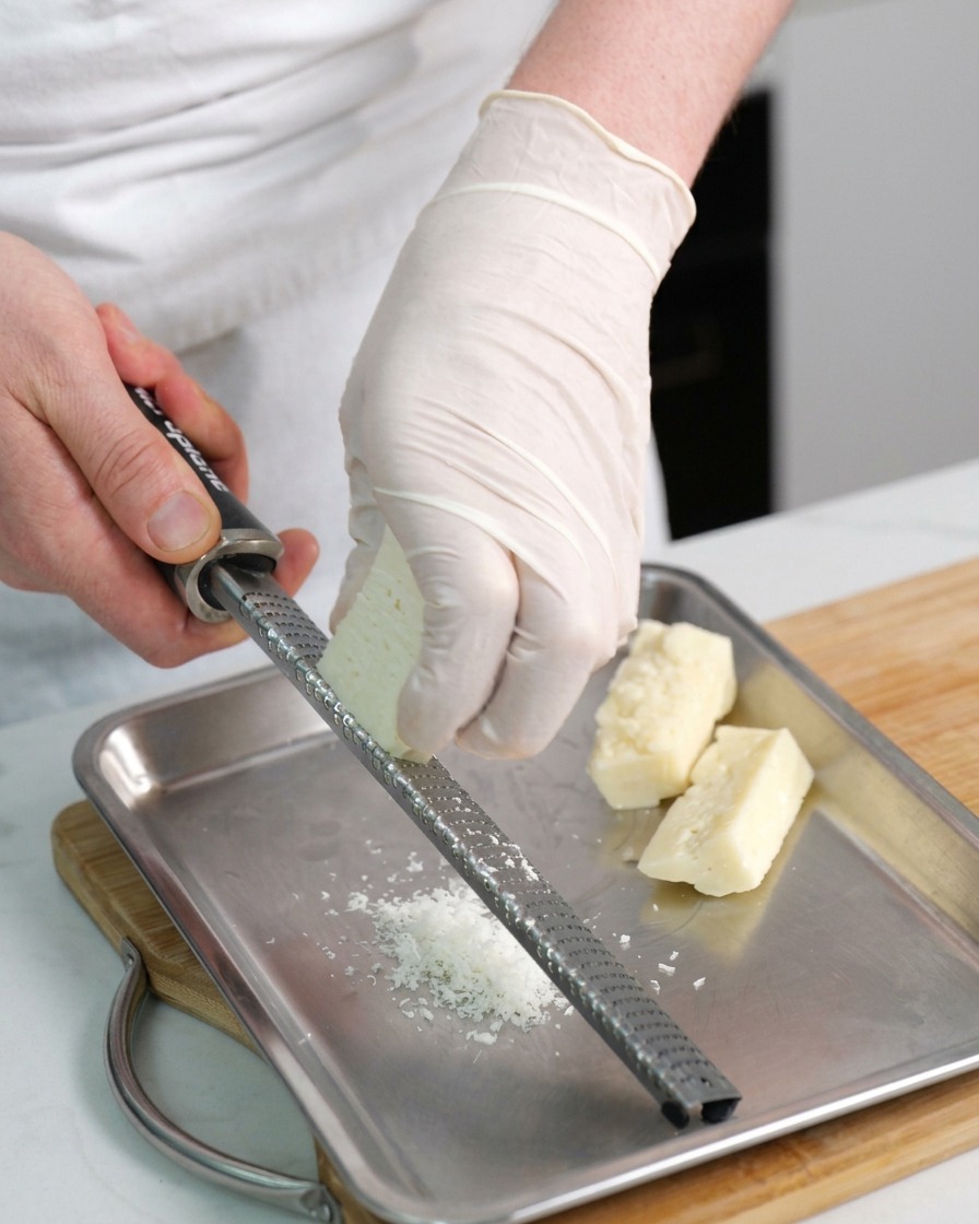 Dłonie w rękawiczkach ścierające kawałek sera Pecorino Romano na tarce nad metalową tacą.