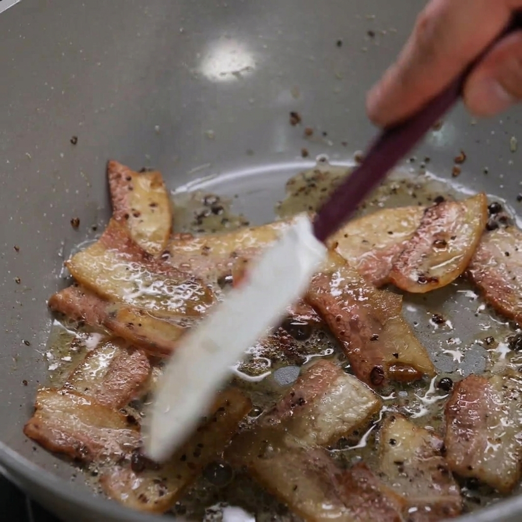 Pokrojone guanciale rumieniące się na patelni z wytapiającym się tłuszczem, mieszane jasną szpatułką.