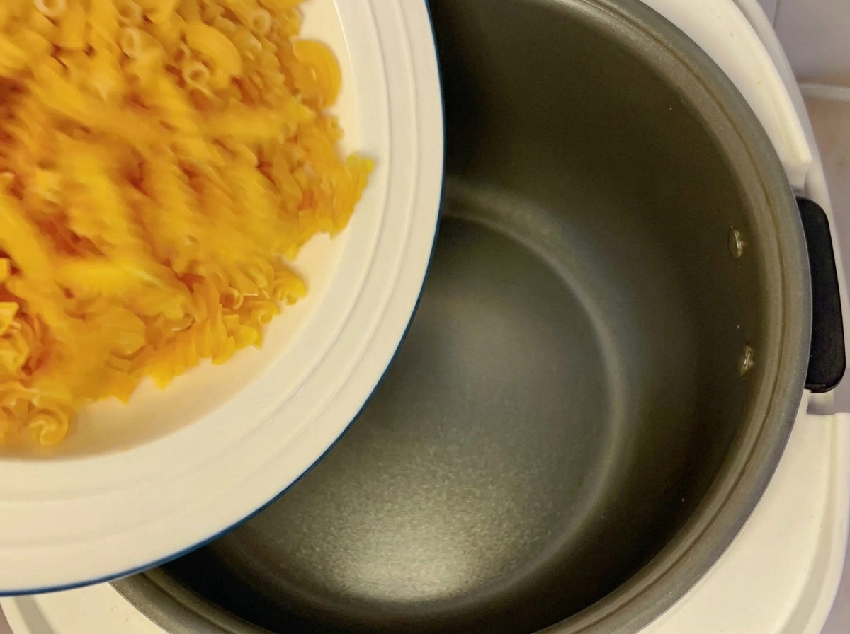 Suchy makaron fusilli wsypywany z białego talerza do ciemnej misy ryżowaru.