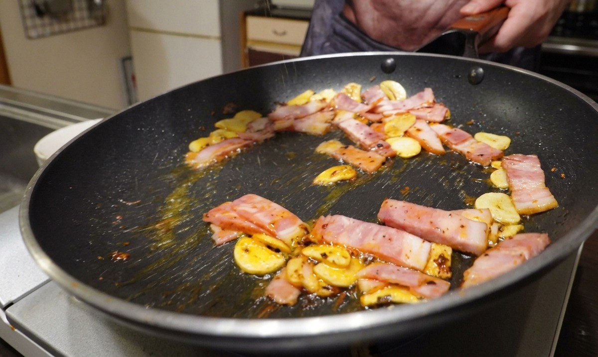 Boczek, czosnek i chili smażące się na patelni, doprawiane czarnym pieprzem.