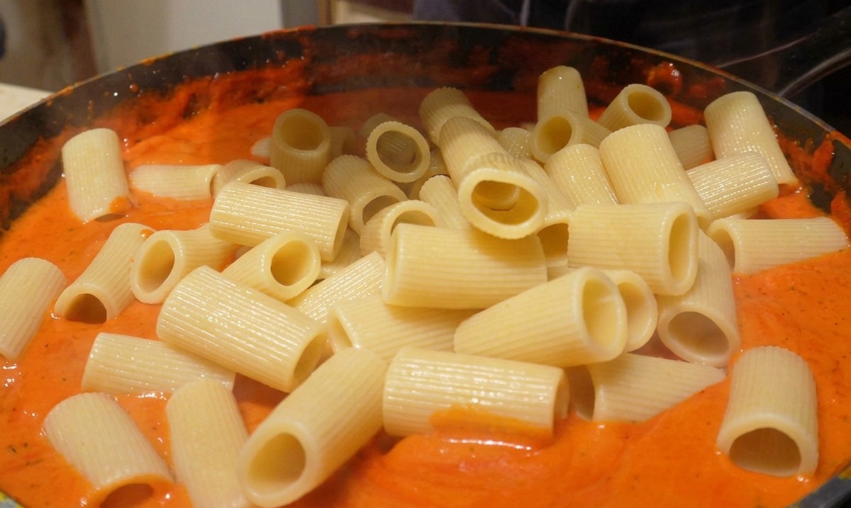 Dodawanie ugotowanego makaronu rigatoni do patelni z gładkim, pomarańczowym sosem.