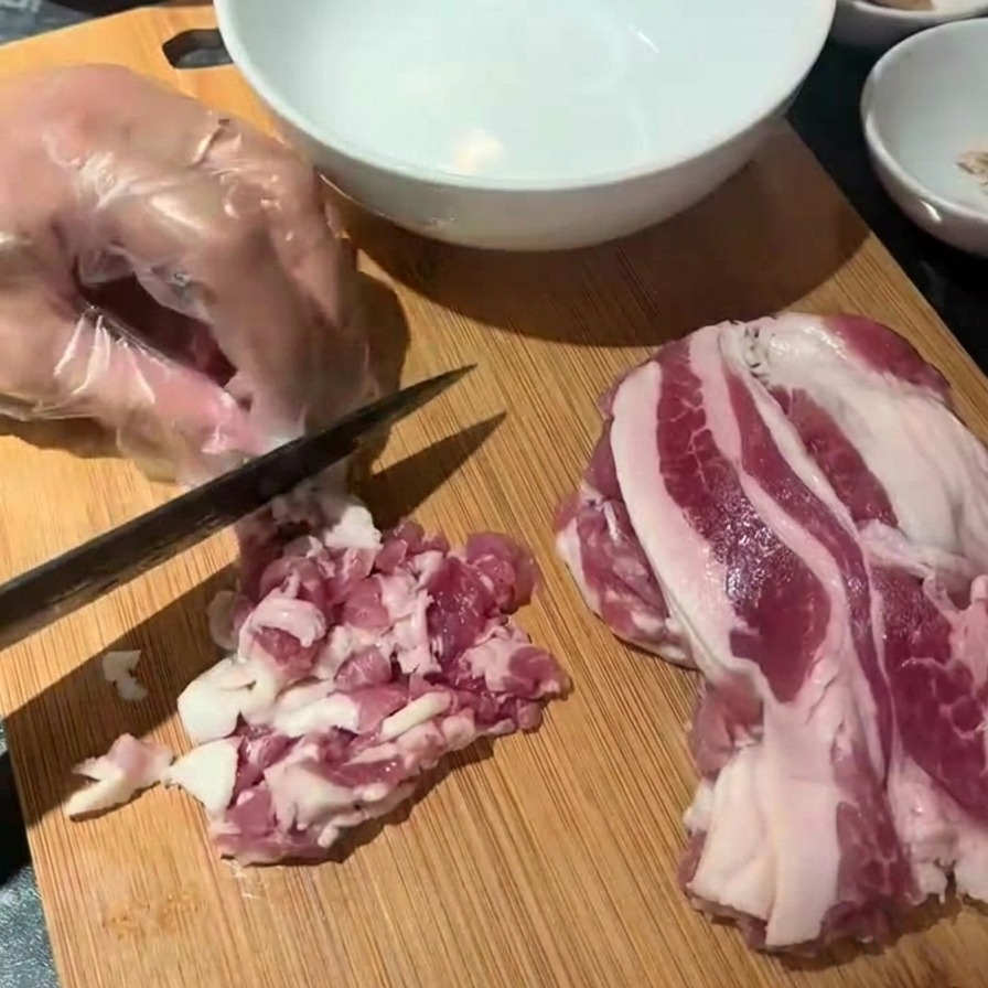 Ręka w rękawiczce używająca czarnego noża do krojenia surowego boczku na małe kawałki na drewnianej desce.