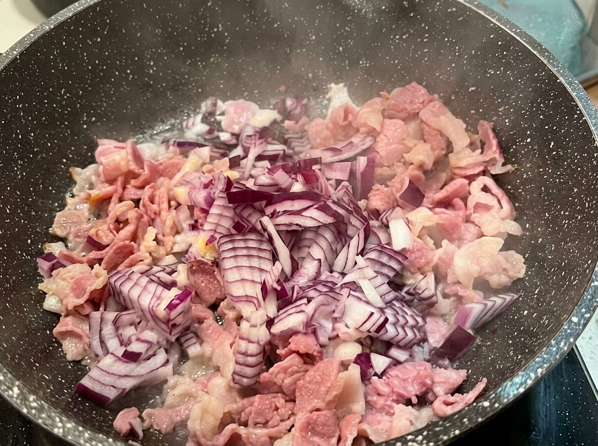Posiekana czerwona cebula na patelni obok podsmażonego boczku.