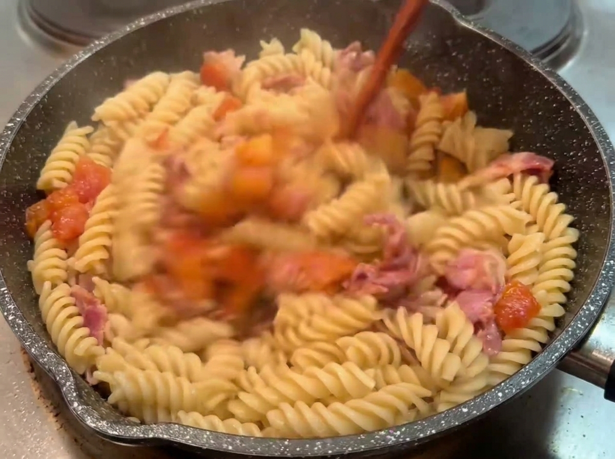 Makaron fusilli mieszany drewnianą łopatką na patelni z gęstym sosem pomidorowo-boczkowym.