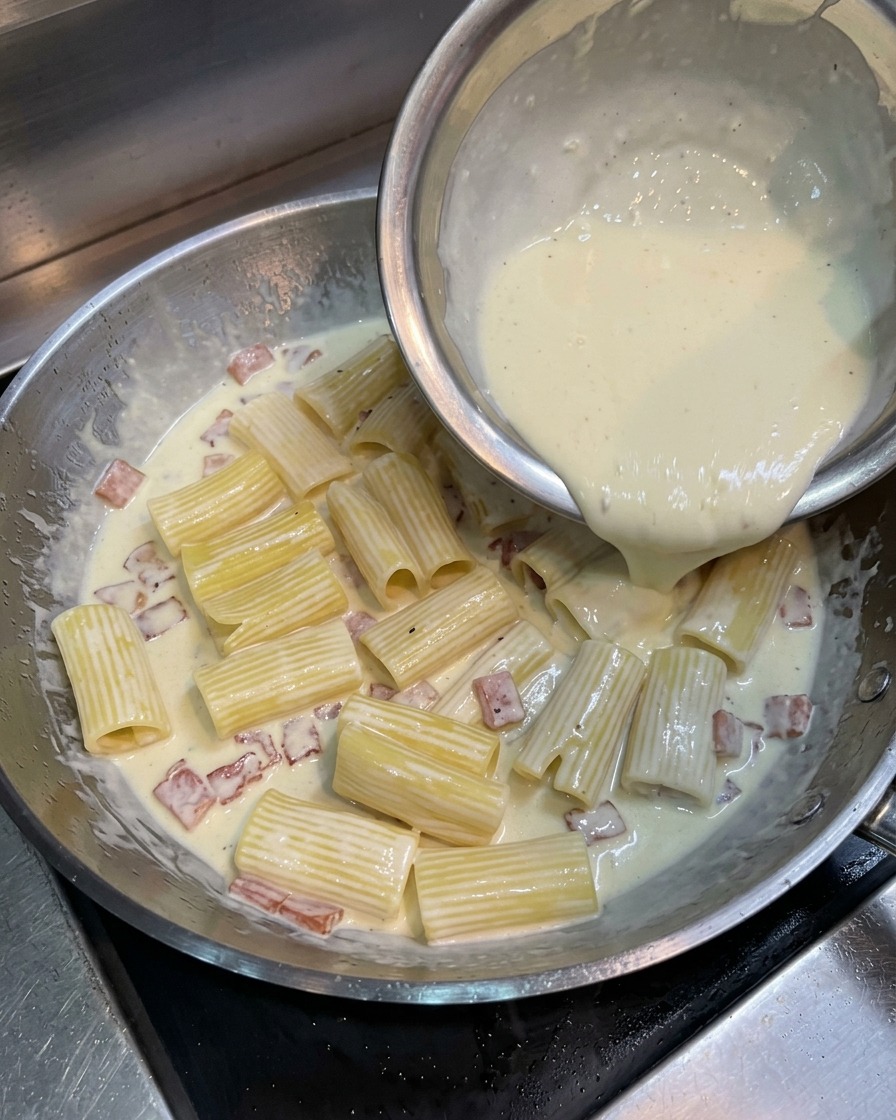 Wlewanie jasnożółtej masy z żółtka, śmietanki i sera na makaron rigatoni z boczkiem na patelni.