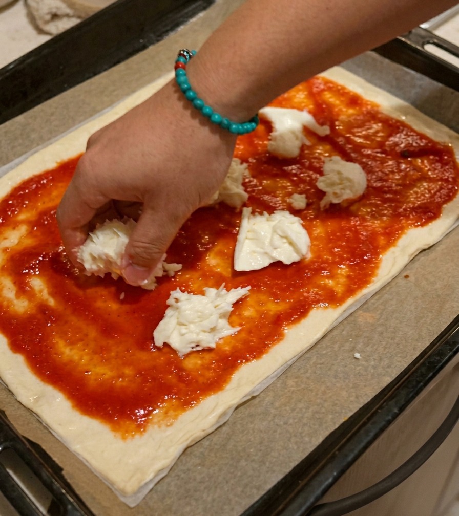 Dłoń układająca nieregularnie urwane kawałki białej mozzarelli na spodzie pizzy pokrytym czerwonym sosem.