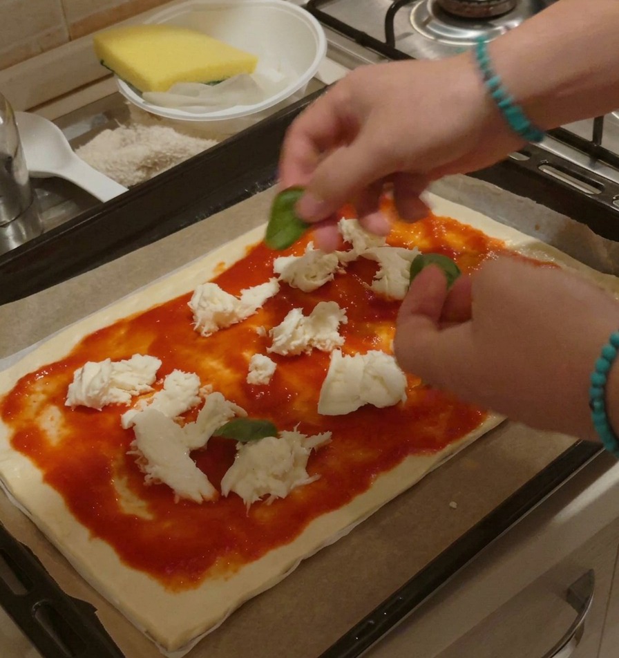 Dłonie ostrożnie układające całe, świeże listki bazylii na surowej pizzy pokrytej sosem i kawałkami mozzarelli.
