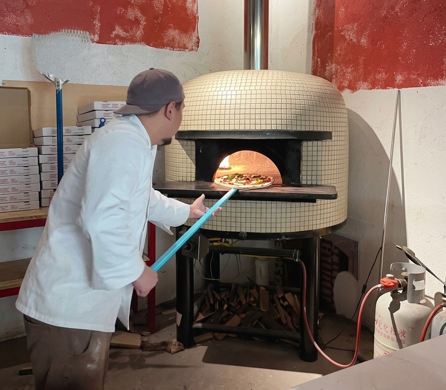 Kucharz wsuwa pizzę do gorącego pieca chlebowego za pomocą niebieskiej łopaty.