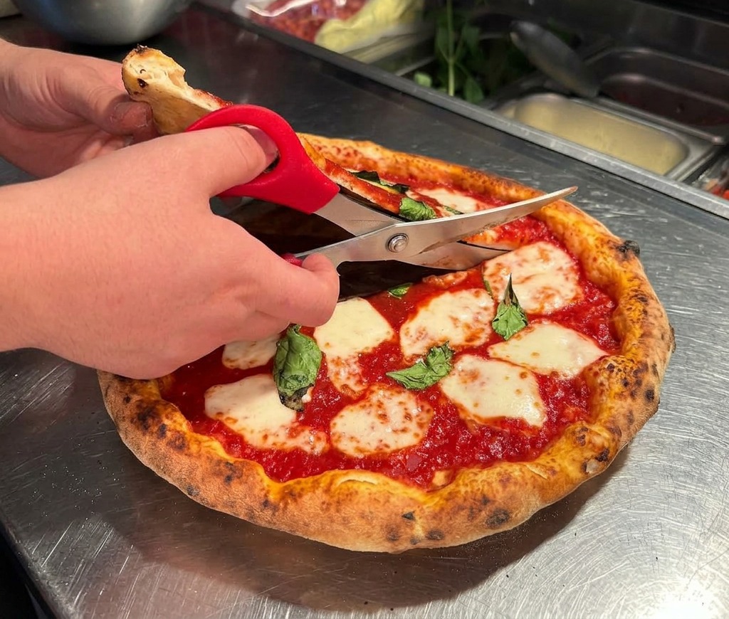 Dłonie używające nożyczek kuchennych do krojenia gorącej pizzy na stalowym blacie.