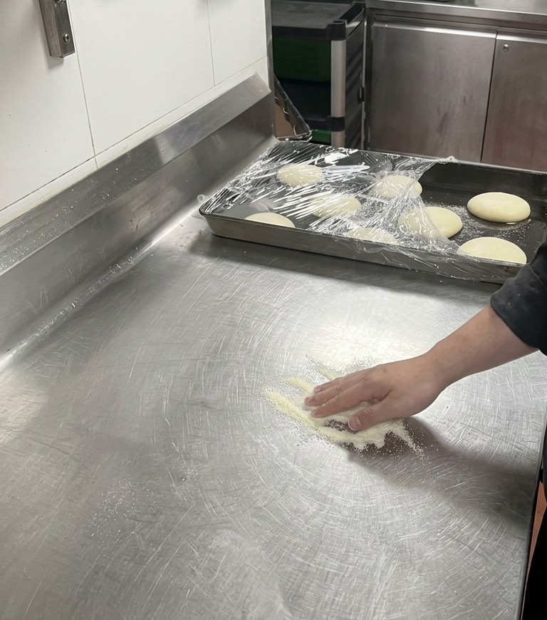 Dłoń kucharza delikatnie dociskająca kulkę ciasta do pizzy do kopca białej mąki na metalowym blacie.