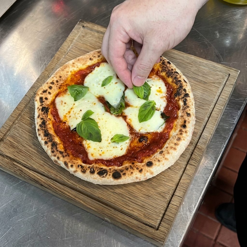 Dłoń układająca świeże liście bazylii na gorącej pizzy Marghericie leżącej na drewnianej desce.