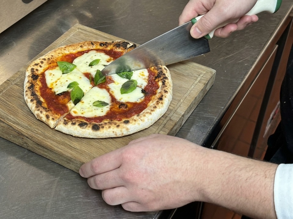 Dłonie krojące pizzę Margheritę na trójkąty dużym nożem kuchennym na drewnianej desce.