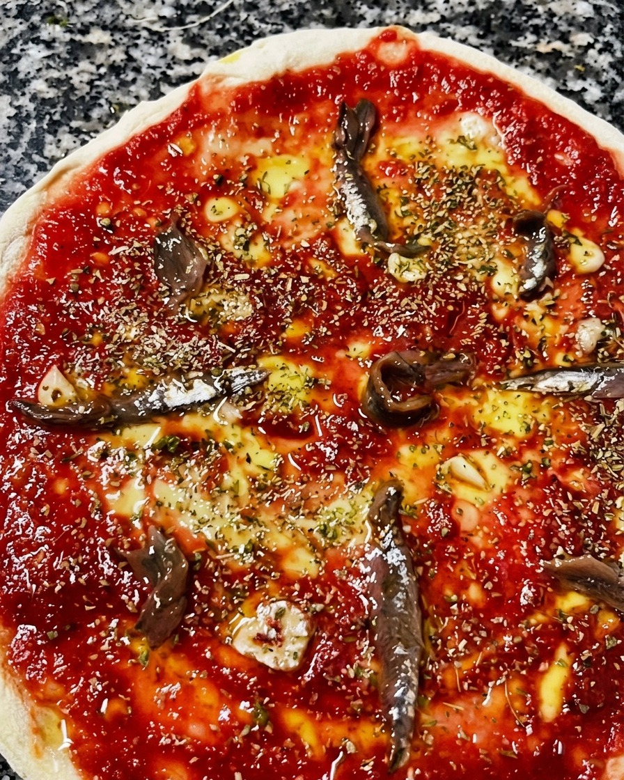 Widok z góry na surową pizzę z sosem, oliwą i oregano, na której rozłożono filety anchois.