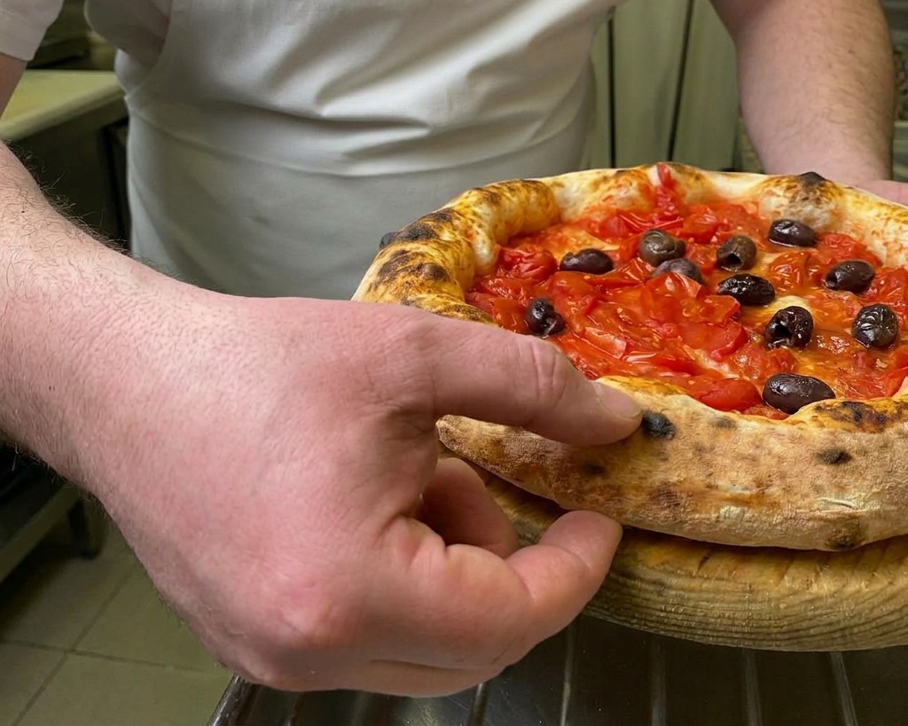 Szef kuchni wciskający palec w mocno wyrośnięty, przypieczony brzeg gorącej, świeżo upieczonej pizzy z pomidorami i oliwkami, aby sprawdzić sprężystość ciasta.