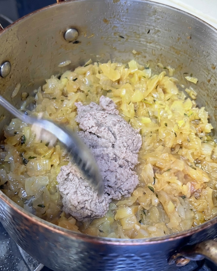 Łyżka szarej pasty rybnej dodawana do garnka z mocno skarmelizowaną cebulą.