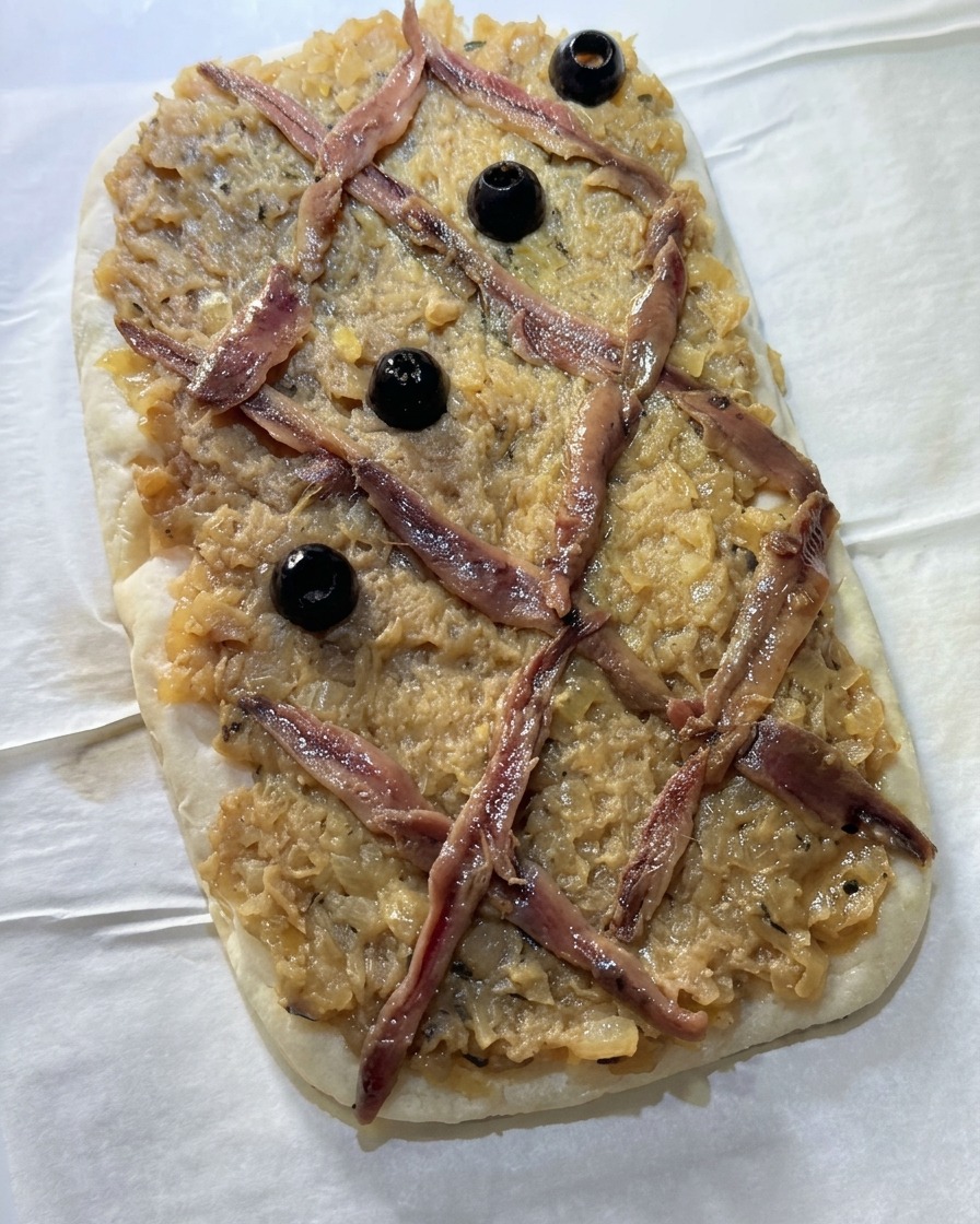 Całe czarne oliwki układane w środku rombów utworzonych przez kratkę z anchois na wierzchu cebulowego placka.