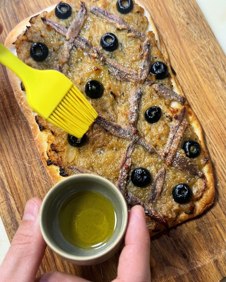 Dłoń używająca żółtego silikonowego pędzelka do smarowania oliwą świeżo upieczonej Pissaladière.